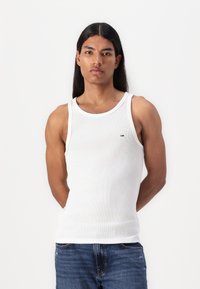 Weißes geripptes Tanktop mit Rundhalsausschnitt und einem kleinen Logodetail, kombiniert mit blauen Jeans aus Denim. Glatte Textur und figurbetontes Design.