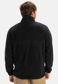 Mustaga fliisjakki kõrge kaelusega, tekstuurse pinna ja lihtsa selja disainiga. Pikad varrukad ja kergelt kõverdatud serv.