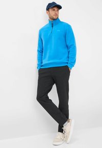 Pullover in pile azzurro chiaro con mezza zip, abbinato a pantaloni grigi scuri. Il modello indossa sneakers color crema e un cappellino scuro. Tessuto complessivamente liscio.