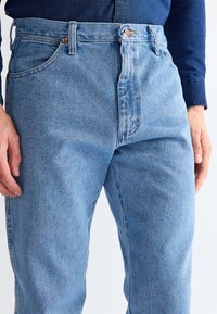 Man wearing light blue denim jeans and a dark blue long-sleeve shirt, hands relaxed by sides against a white background.