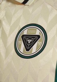 Sirkulært merke på kremfarget sportstrøye med geometrisk mønster, med romertallene XXIII og en sentral stilisert ugle inne i en trekant.