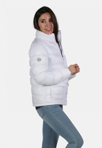 Doudoune blanche avec col haut, fermeture éclair et poches intérieures. Présente un patch logo rond sur le bras gauche. Portée avec un jean bleu clair.