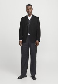 Zwarte blazer met notch-revers, drie knopen en opgestikte zakken, gecombineerd met een donkere vest en bijpassende broek, afgerond met zwarte schoenen.