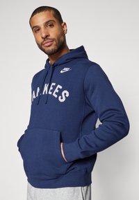 Navy zila džemperis ar kabatu, balta "Yankees" uzraksts un mazs balts Nike logo uz krūšu. Mīksta auduma, ribota aproces un apakšmalas.