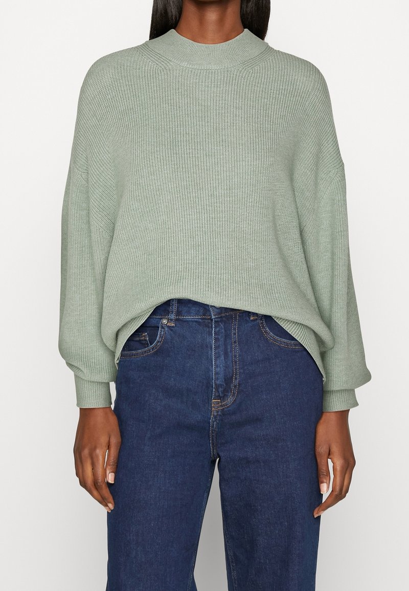 Femme portant un pull ample vert clair à côtes avec manches longues et un jean bleu foncé taille haute.