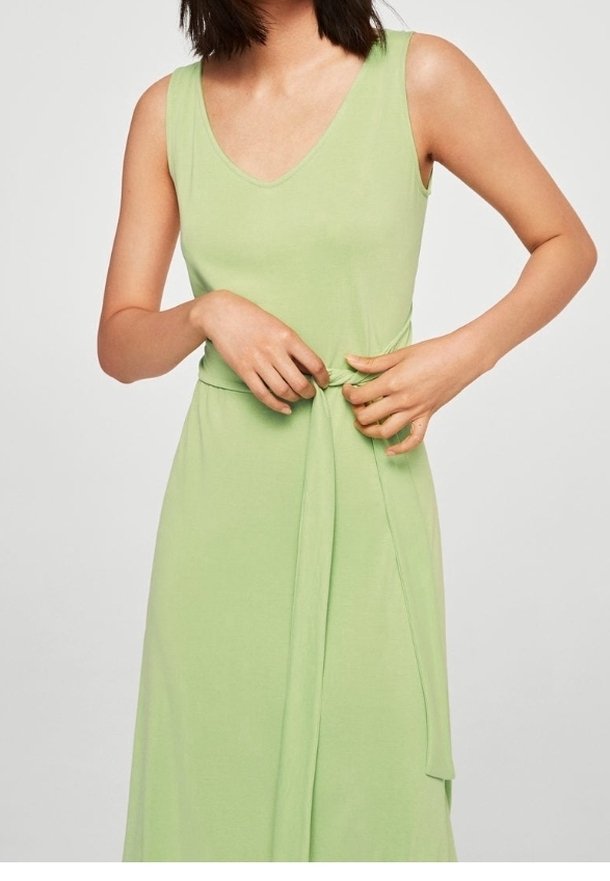 Femme portant une robe vert clair sans manches nouant une ceinture en tissu autour de la taille sur fond uni.