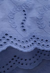 Ljusblått tyg med intrikata broderier i blommönster och cirkulära utskärningar längs kanten, vilket tillför textur och detalj.