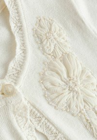 Cardigan en tricot crème avec une broderie florale texturée comportant des pétales superposés et de petites perles, complété par un bouton décoratif.