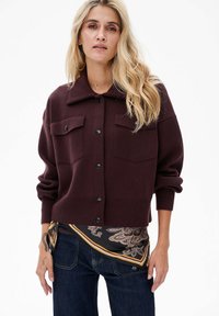 Bordowy kardigan z kołnierzem, dwoma kieszeniami na piersi i guzikami z przodu. Połączony z czarnym szalikiem w wzory i ciemnymi dżinsami.