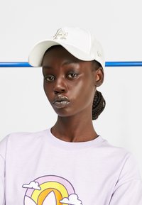 Gorra blanca de béisbol con el logotipo bordado "LA", combinada con una camisa lilac clara que presenta un diseño de arcoíris colorido. Textura de tela suave.