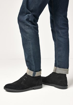Person trägt dunkelblaue hochgekrempelte Jeans und schwarze Wildleder-Schnürstiefeletten und steht auf einer weißen Fläche.