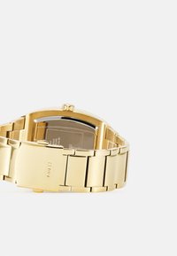 Montre-bracelet dorée avec un boîtier rectangulaire et un bracelet à maillons, présentant une finition polie et un logo de la marque gravé sur le fermoir.