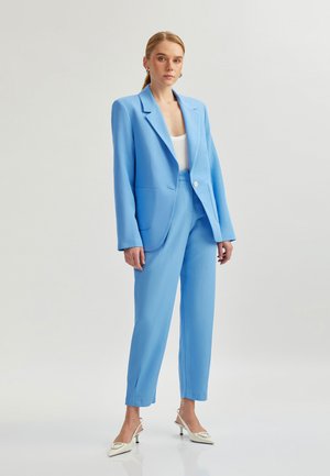 Lichtblauw op maat gemaakt kostuum met een blazer met notched revers en twee voorzakken, gecombineerd met bijpassende toegesneden broek. Witte top en hakken.