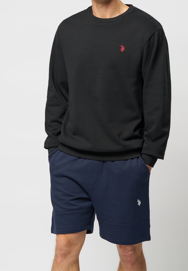Homme portant un sweat-shirt noir à manches longues avec un logo rouge et un short bleu marine avec un logo blanc, mains dans les poches, sur fond clair.