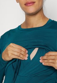 Teal graviditetstopp med ett mjukt, elastiskt tyg, med en crossover-design och diskret öppning för amning.