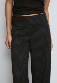 Pantalones de raya diplomática negros con una cinturilla ancha, que presentan líneas plateadas sutiles y un corte recto, fabricados en un material suave.