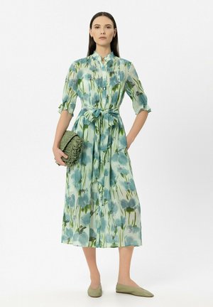 Frau in einem blau-grünen Midikleid mit Blumenmuster und halblangen Ärmeln, mit gebundener Taille, die eine grüne, strukturierte Clutch hält und grüne Ballerinas trägt.