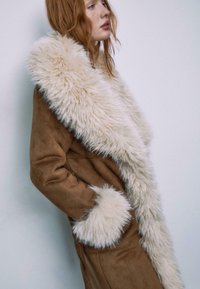 Suedesjakke i brun med en fyldig, lysebeige faux fur-krave og manchetter. Har et strømlinet design og sidelommer.