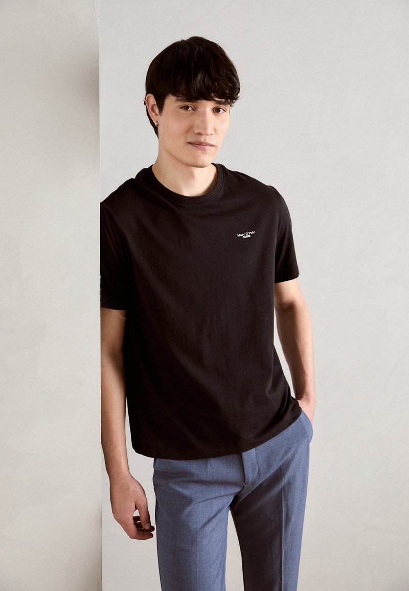 Zwarte katoenen t-shirt met een ronde hals en een klein wit logo. G gedragen met lichtblauwe op maat gemaakte broek, tegen een minimalistische achtergrond.