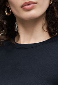 Primer plano de la parte inferior del rostro y cuello de una persona, que lleva pendientes de aro dorados y una camiseta negra acanalada con cuello redondo.