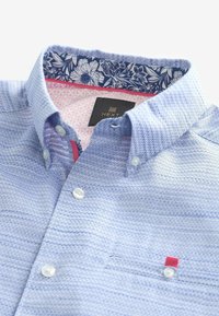 Chemise bleu clair texturée avec boutons blancs, motif floral à l'intérieur du col, doublure intérieure rose et petite languette rose sur la poche poitrine.