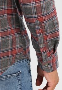 Camisa de franela a cuadros en rojo y gris con un acabado texturizado, que presenta puños abotonados y un dobladillo curvado, mostrada junto a unos pantalones vaqueros azules.