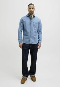 Camicia di jeans azzurri con colletto e taschino anteriore, abbinata a jeans blu scuro e mocassini color cuoio. Il modello indossa una sciarpa marrone attorno al collo.