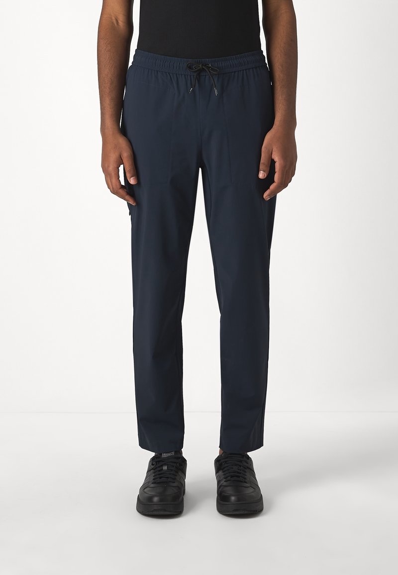 Homme portant un pantalon bleu marine à cordon de serrage et des baskets noires, debout devant un fond blanc uni.