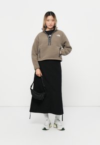 Lys brun fleece-pullover med halvmeter glidelås, to frontlommer og svarte detaljer, kombinert med en lang svart skjørt og hvite sneakers.