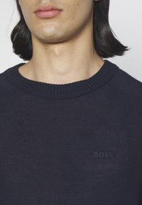 Gros plan d'une personne portant un pull bleu marine foncé à col rond avec "BOSS" brodé sur la poitrine.