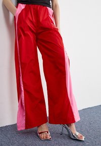 Pantalons larges rouges et roses en tissu léger et brillant, avec des poches latérales et des panneaux contrastants le long des coutures.