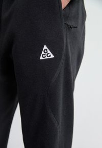 Schwarze Fleece-Hosen mit einem weißen gestickten Dreieckslogo. Glatte Textur mit schmalem Schnitt und Seitentaschen. Verstellbarer Kordelzugbund.