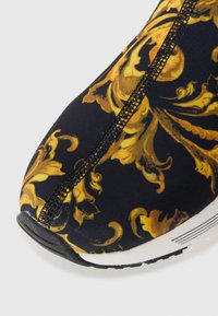 Scarpa in tessuto nero con un motivo barocco dorato, superficie texturizzata e punta rotonda, con suola bianca e cuciture a contrasto.