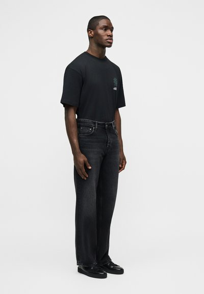Άνδρας που στέκεται κοιτάζοντας ελαφρώς προς τα δεξιά, φοράει μαύρο t-shirt με σχέδιο, σκούρο γκρι τζιν και μαύρα παπούτσια σε φωτεινό απλό φόντο.