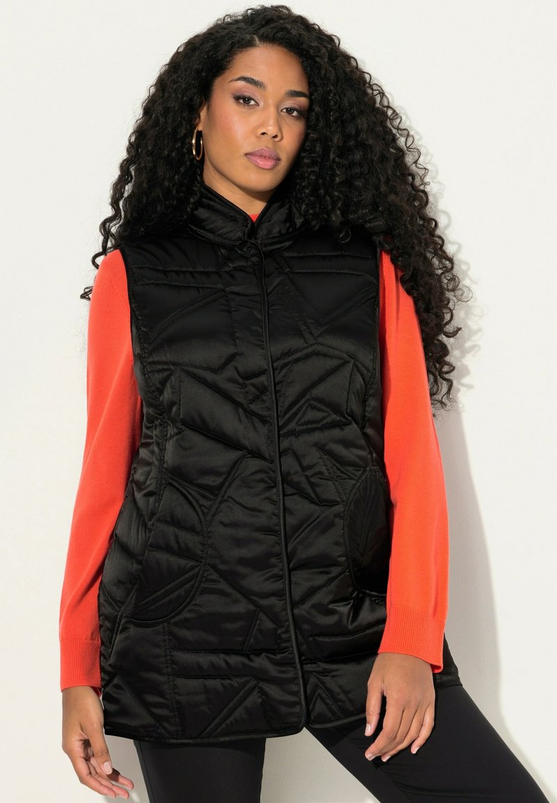 Černá prošívaná vesta s vysokým límcem, předním zipem a bočními kapsami, nošená přes jasně oranžové dlouhé rukávy. Texturovaná tkanina s lesklým povrchem.