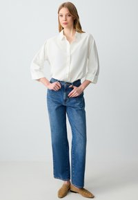 Witte button-up blouse met een gestructureerde afwerking, gecombineerd met hoge blauwe spijkerbroek. Model draagt tan loafers met metalen accenten.