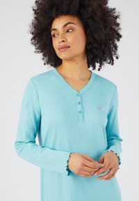 Donna con capelli ricci che indossa un top a maniche lunghe ribbed di colore azzurro chiaro con bottoni, che guarda leggermente di lato su uno sfondo neutro.