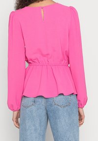 Maglietta rosa a maniche lunghe con vita elastica e design a peplo, caratterizzata da un dettaglio a buco chiave sulla schiena, abbinata a jeans in denim azzurro chiaro.