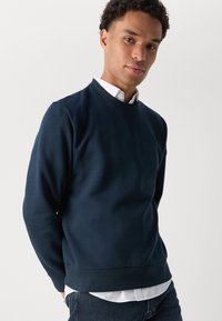 Felpa blu navy realizzata in tessuto liscio, con un collo a girocolo a coste e un orlo con polsini. Indossata sopra una camicia bianca con colletto.