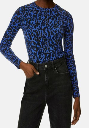 Frau trägt ein langärmeliges Oberteil mit blau-schwarzem Leopardenmuster, das in eine hoch taillierte schwarze Jeans gesteckt ist, mit einer Hand in der Tasche.