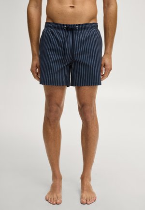 Mann trägt marineblaue Badeshorts mit vertikalen hellblauen Streifen, steht barfuß vor einem einfarbigen Hintergrund.