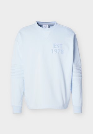 Deux personnes portant des sweat-shirts bleu clair avec l'inscription "EST 1978", des pantalons larges brun, et des chaussures noires ; l'une tient un sac noir.