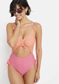 Enodelni kopalni kostim z naborki v koralni in roza barvi, z nastavljivimi bočnimi trakovi in izrezom v srednjem delu. Gladka tekstura tkanine.