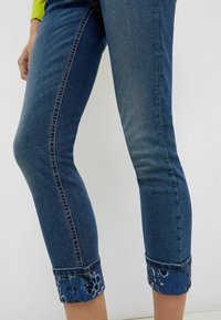 Jeans de denim azul recortados con puños de patrón floral doblados, llevados por una persona con una manga amarilla visible.