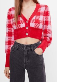 Cardigan corto a quadri rossi e rosa con scollo a V profondo, tessuto strutturato e tre bottoni trasparenti; abbinato a jeans neri a vita alta.