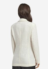 Biały żakiet z teksturowanym wzorem w pionowe linie i małymi, błyszczącymi akcentami. Posiada klasyczny kołnierz i długie rękawy, dopasowany krój.