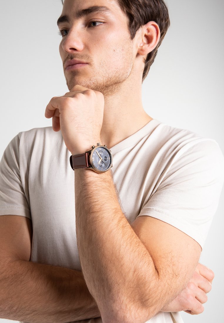 Joven con una camiseta blanca, con los brazos cruzados, apoyando el mentón en el puño, que lleva un reloj de pulsera con correa de cuero marrón y esfera azul.