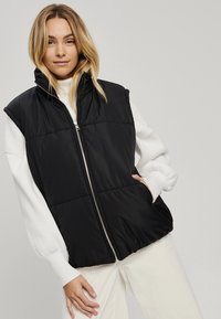 Juodas paminkštintas be rankovių liemenė su aukštu apykakle ir priekiniais užtrauktukais, turinti siuvinėtą dizainą ir šonines kišenes. Dėvima ant baltos megztinio.