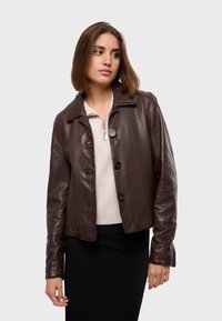 Veste en cuir marron avec un col, fermeture à boutons sur le devant et manches longues. Superposée à un haut zippé crème et une jupe noire.