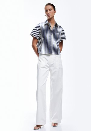 Striped short-sleeve button-up shirt in navy and white, paired with wide-leg white pants. Model stands with hands on hips, showcasing relaxed fit.